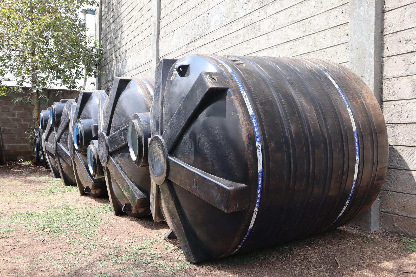A line of large black horizontal Jumbo tanks resting on their sides along a stone wall, with open inlet collars facing the camera.