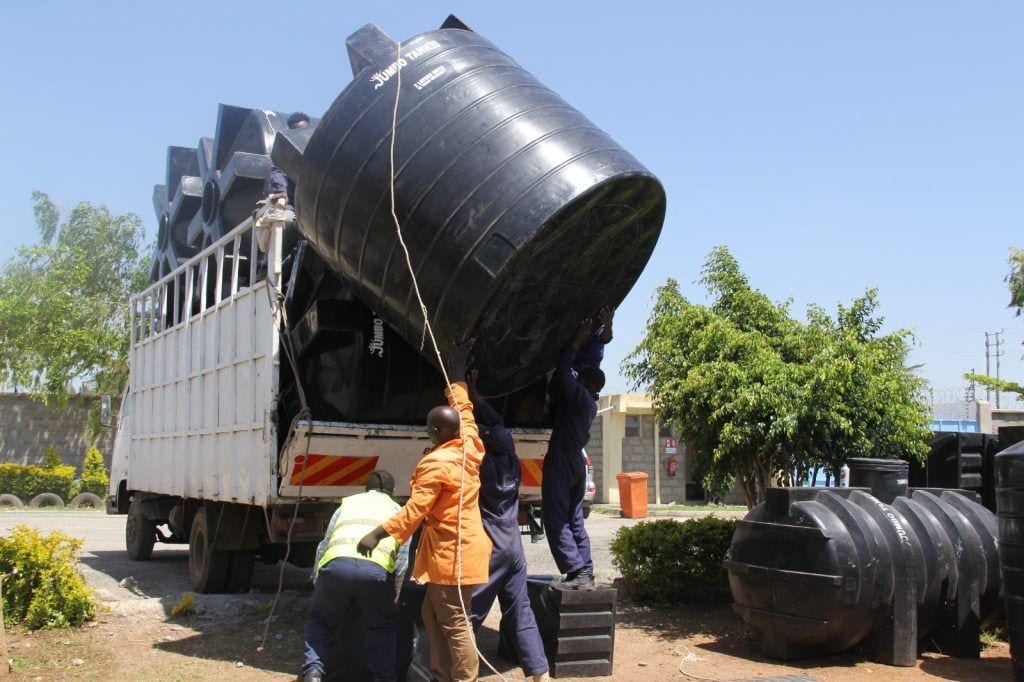 Four Jumbo Tanks crew in overalls and hi-vis roping a large black JUMBO TANKS cylindrical tank up onto a lorry bed at the yard, with more tanks already loaded behind them.